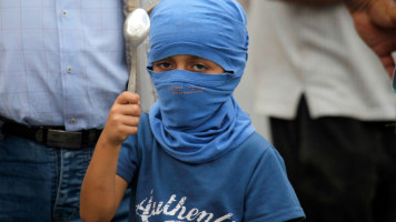 A Palestinian boy with covered face holds a spoon as a symbol during the demonstration. Masked Palestinians protest in solidarity with six Palestinians who escaped from the Israeli Gilboa prison on September 6th through a tunnel dug with spoons under the sink [Getty Images]