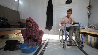 A woman washes clothes in a plastic tub and a girl in a wheelchair watches