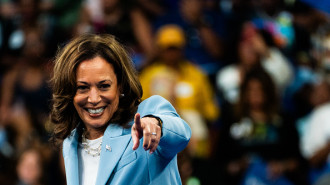 Woman wearing a white top and pale blue blazer pointing towards camera