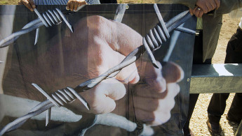 Palestinians holding a banner which has a person holding prison bars with barbed wire overlaid