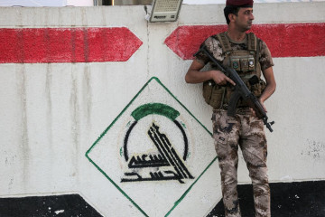 A fighter in Iraq's Popular Mobilisation Forces stands guard next to a wall showing the group's logo outside their headquarters in the capital Baghdad on 13 June, 2021. [Getty]