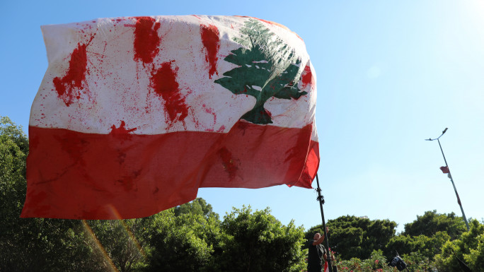 A man waves a Lebanese flag with fake blood smeared on it at a protest to commemorate the third anniversary of the Beirut port explosion. [William Christou - TNA] 