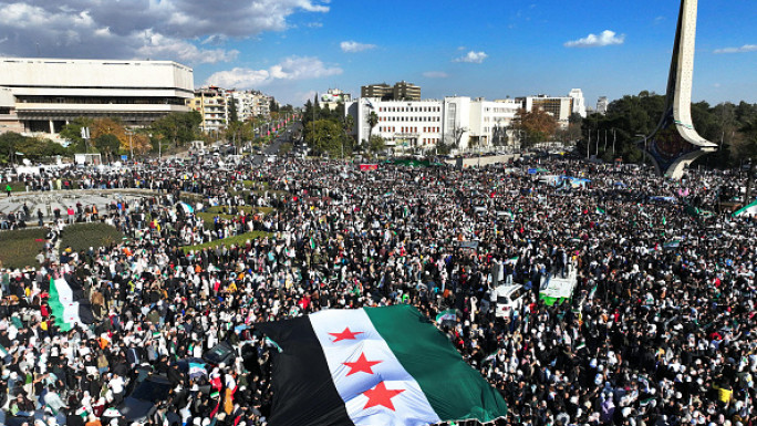 An areal photo shows crowds of Syrians raising a giant independence-era flag, as they celebrate the fall of Bashar al-Assad's rule earlier this week at the central Umayyad Square in Damascus on December 13, 2024. (OMAR HAJ KADOUR/AFP via Getty)