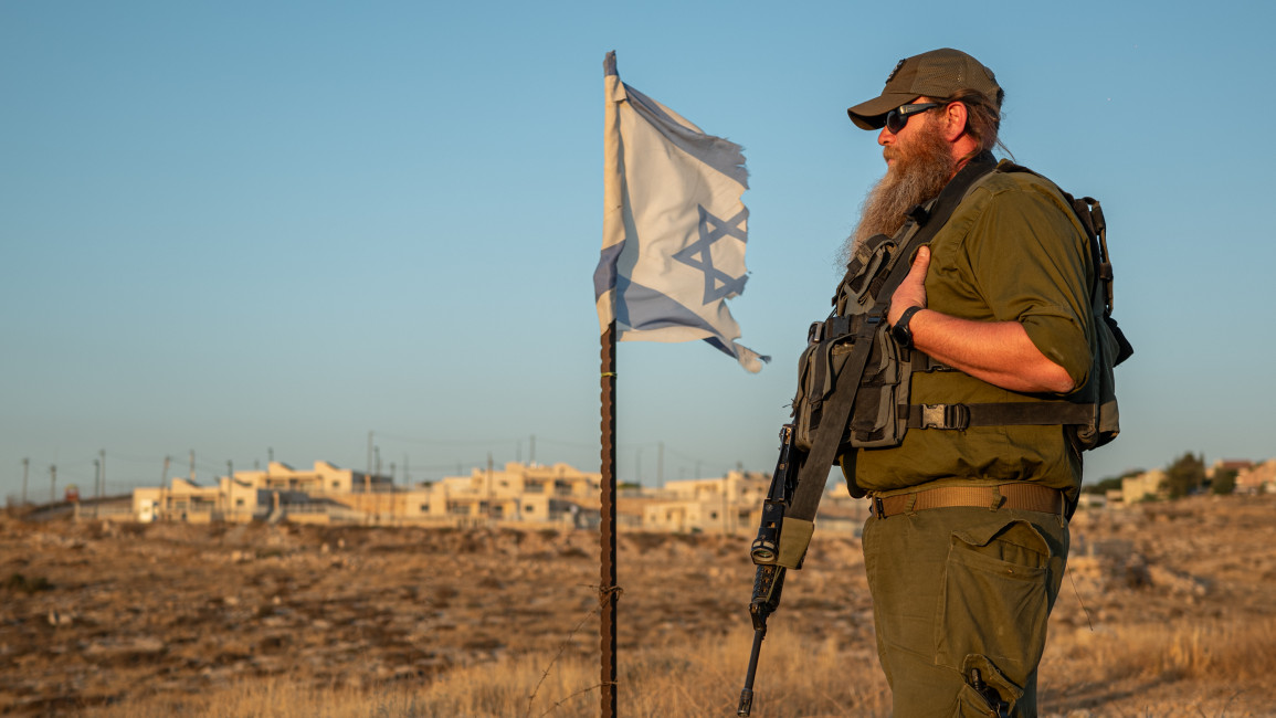 Man in military fatigues with a gun next to an Israeli flag standing in a field with buildings in background