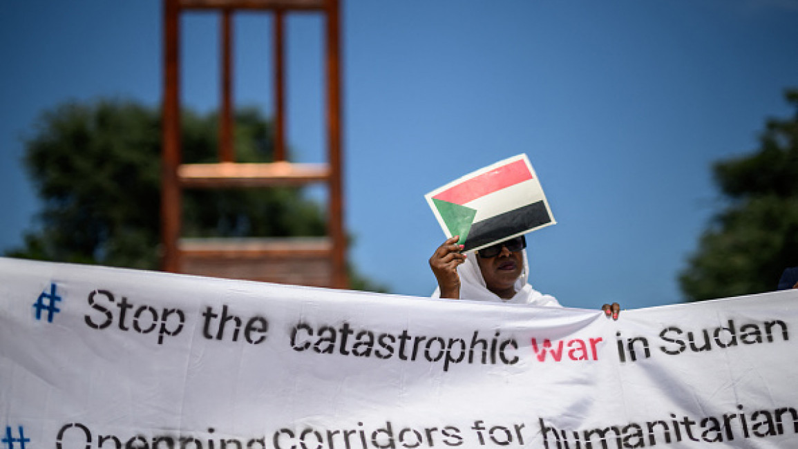 A woman protecting her face with a cardboard bearing the Sudanese flag as she holds a banner next to the monumental wood sculpture "Broken Chair" (L) during a demonstration on the opening day of Sudan ceasefire talks, in Geneva, on August 14, 2024. US-sponsored talks on agreeing a ceasefire in the devastating conflict in Sudan kicked off in Switzerland, despite the Sudanese government staying away.